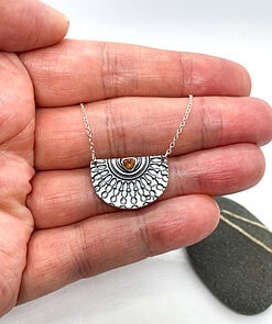 A hand holding a silver semicircle pendant with a detailed pattern and an amber gemstone in the center.