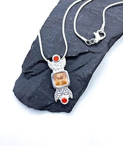 Silver necklace featuring a pendant with an orange square gemstone and two red round gems, displayed on dark stone.