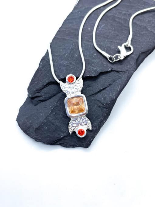 Silver necklace featuring a pendant with an orange square gemstone and two red round gems, displayed on dark stone.