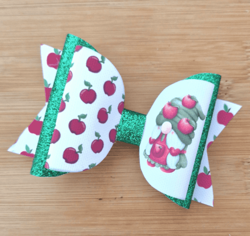 A colorful hair bow with an apple pattern and a gnome design on a wooden background. The bow has green glitter accents.