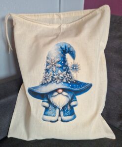 A beige drawstring bag featuring a gnome in a blue hat adorned with snowflakes and crystals, sitting on a gray cushion.