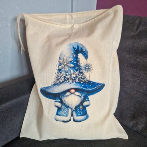 A beige drawstring bag featuring a gnome in a blue hat adorned with snowflakes and crystals, sitting on a gray cushion.