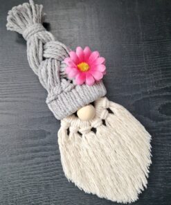 Macrame gnome with a braided gray hat, pink flower accent, wooden nose, and white fringed beard on a dark surface.