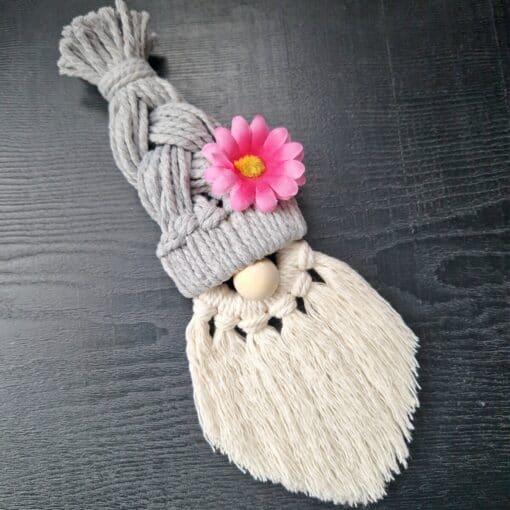 Macrame gnome with a braided gray hat, pink flower accent, wooden nose, and white fringed beard on a dark surface.