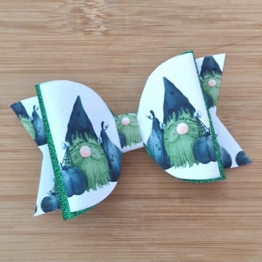 White hair bow with green and black gnome pattern, accented with green glitter edges, on a wooden background.