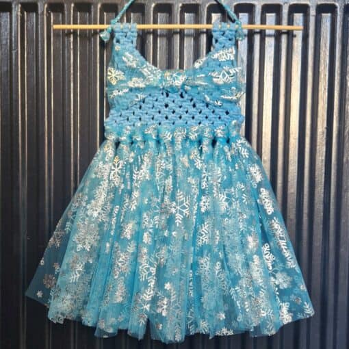 Blue and white crocheted dress with a snowflake pattern, hanging on a wooden rod against a black corrugated background.