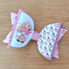 Pink and white decorative bow with glitter and peach patterns, featuring an illustration of a character on a wooden background.