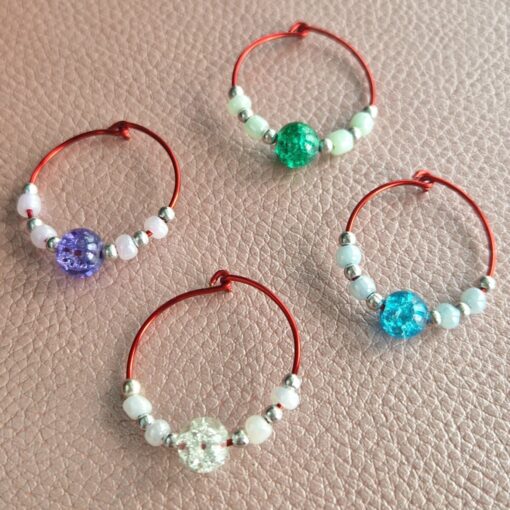 Four red wire wine glass charms with different colored beads: purple, green, blue, and clear, placed on a textured surface.