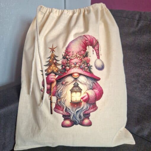 Cloth drawstring bag with a festive gnome holding a lantern and small decorated Christmas tree, sitting on a gray surface.