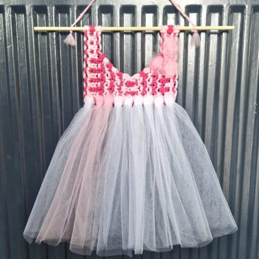 Handmade pink and white crochet top dress with a tutu skirt, hanging on a wooden rod against a corrugated metal background.