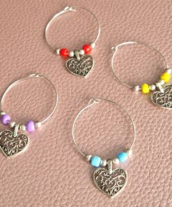 Four wine glass charms with metal heart pendants and colorful beads, displayed on a textured pink surface.