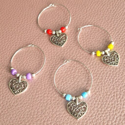 Four wine glass charms with metal heart pendants and colorful beads, displayed on a textured pink surface.