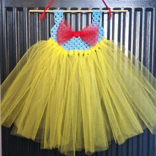 Handmade yellow tulle dress with a blue crocheted bodice and red bow, hanging on a black background.