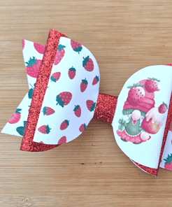 Strawberry-themed hair bow with red glitter and white ribbon featuring strawberries and gnomes, placed on a wooden surface.