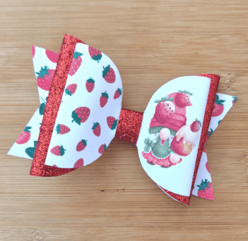 Strawberry-themed hair bow with red glitter and white ribbon featuring strawberries and gnomes, placed on a wooden surface.