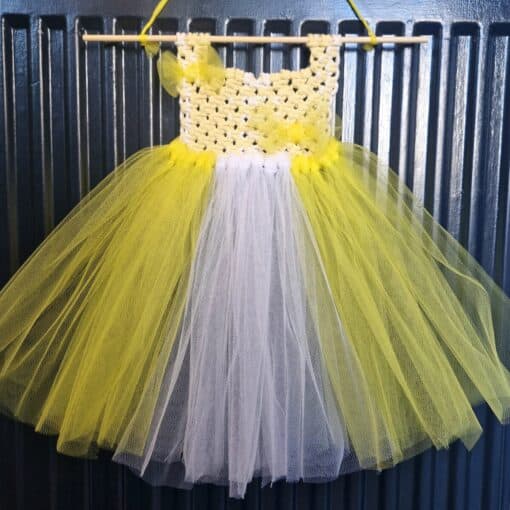 Yellow and white crocheted and tulle tutu dress hanging on a wooden dowel against a dark metallic background.