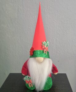 A gnome with a long white beard, wearing a tall red hat and green glittery accents, sits indoors against a textured wall.