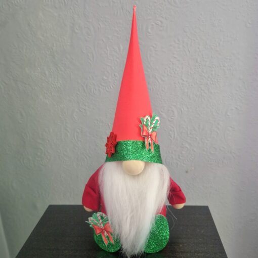 A gnome with a long white beard, wearing a tall red hat and green glittery accents, sits indoors against a textured wall.