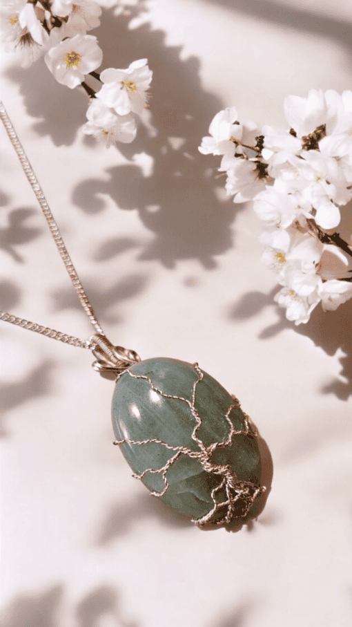 Silver tree of life pendant on green gemstone, surrounded by white cherry blossoms against a soft, shadowed background.