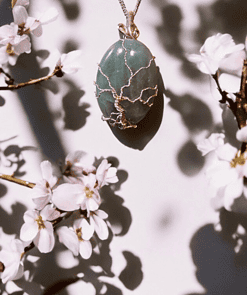 Pendant with a green stone and wire tree design, hanging on a silver chain, surrounded by white blooming flowers.