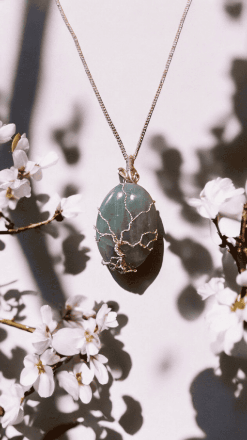 Pendant with a green stone and wire tree design, hanging on a silver chain, surrounded by white blooming flowers.