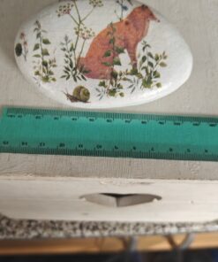 Oval stone painted with a red fox and foliage sits next to a green ruler on a white wooden surface.