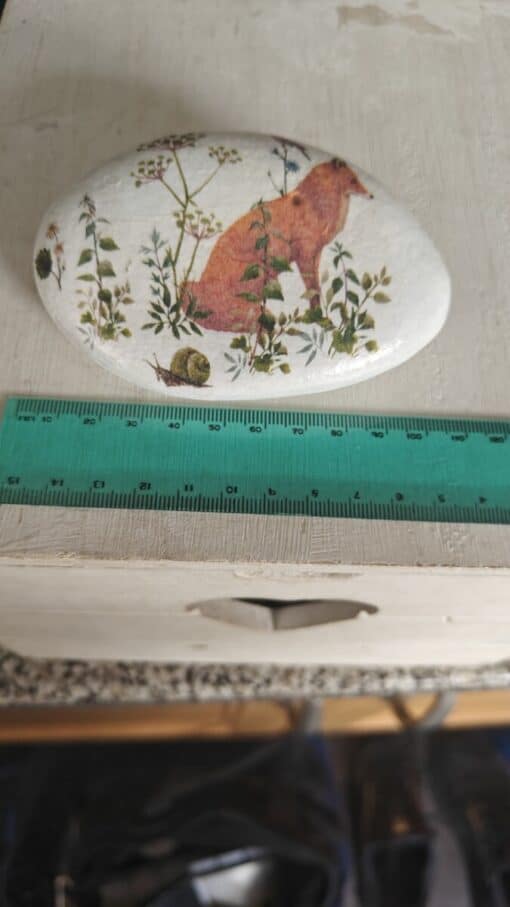 Oval stone painted with a red fox and foliage sits next to a green ruler on a white wooden surface.