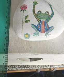 Decorative rock painted with a colorful frog and flowers next to a green ruler on a wooden surface.