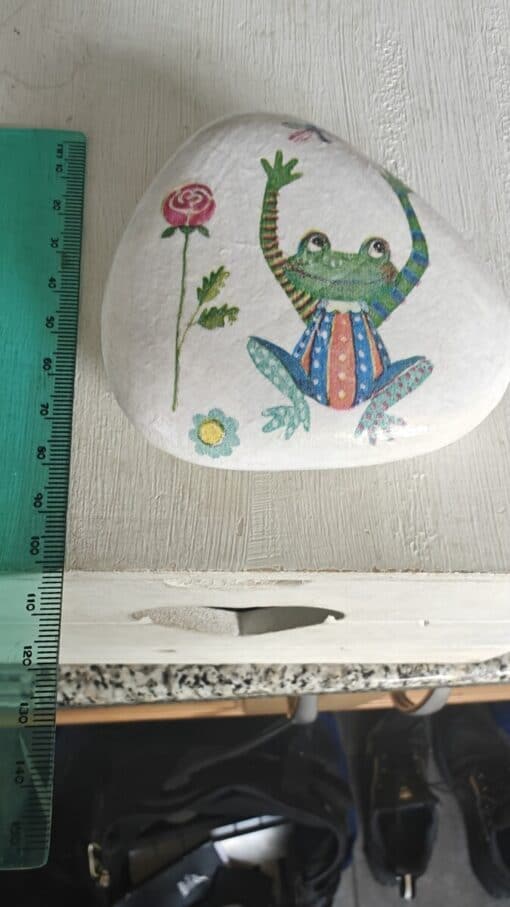 Decorative rock painted with a colorful frog and flowers next to a green ruler on a wooden surface.