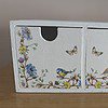 Small white wooden cabinet with two drawers, decorated with floral patterns, birds, and butterflies, placed on a wooden surface.