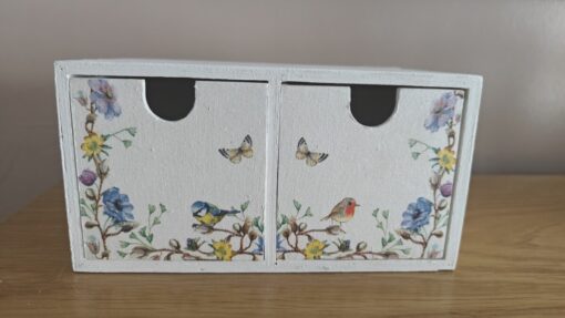 Small white wooden cabinet with two drawers, decorated with floral patterns, birds, and butterflies, placed on a wooden surface.