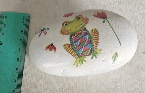 A painted white rock with a colorful frog design, flowers, a dragonfly, and a ladybug next to a green ruler.