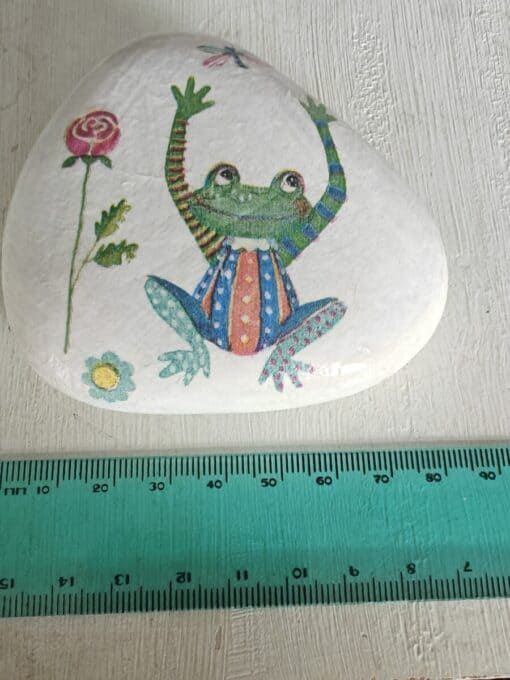 Painted stone with a colorful frog in a striped and polka-dotted outfit, next to a 15 cm green measuring ruler.