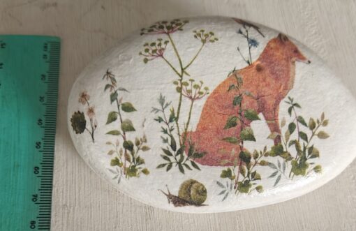 A painted rock with a fox, plants, and a snail, placed next to a green ruler for scale.