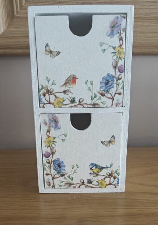 White wooden box with two drawers decorated with floral patterns, birds, and butterflies, placed on a wooden surface.