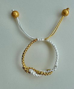 A braided bracelet with gold and white threads, adjustable knots, and gold bead accents on a light background.