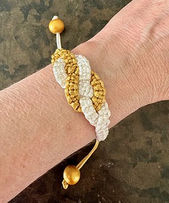 Crocheted bracelet with gold and white intertwined design, worn on a wrist, featuring a wooden bead toggle closure.