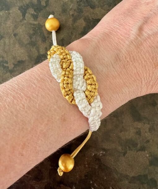 Crocheted bracelet with gold and white intertwined design, worn on a wrist, featuring a wooden bead toggle closure.