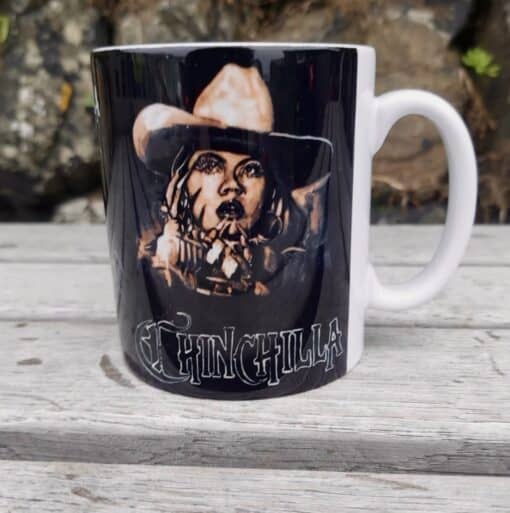 Black mug on wooden table featuring art of a cowboy with "Chinchilla" text.