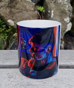 A ceramic mug featuring a vibrant, colorful print of a person, placed on a wooden surface with rocks and greenery in the background.