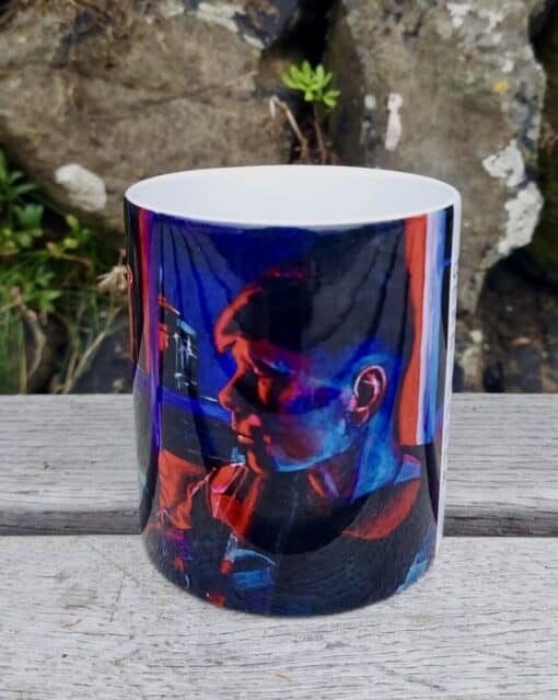 A ceramic mug featuring a vibrant, colorful print of a person, placed on a wooden surface with rocks and greenery in the background.