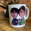 White mug with Oasis cartoon illustrations of two men, one holding a guitar, on a wooden counter beside a vintage kettle.