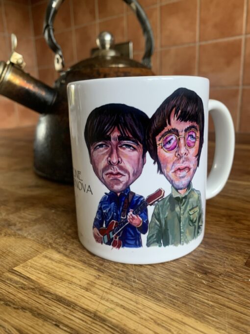 White mug with Oasis cartoon illustrations of two men, one holding a guitar, on a wooden counter beside a vintage kettle.