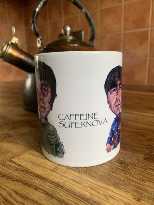 White mug with Oasis caricature illustrations and the text "Caffeine Supernova" on a wooden surface near a vintage kettle.