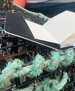 Open UK notebook on a weathered fishing net, showcasing blank pages and a black cover.