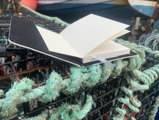 Open UK notebook on a weathered fishing net, showcasing blank pages and a black cover.