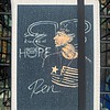 A fabric-covered notebook with a portrait and text: "Some people know me as Hope. Ren." A black elastic band secures it.