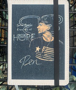 A fabric-covered notebook with a portrait and text: "Some people know me as Hope. Ren." A black elastic band secures it.