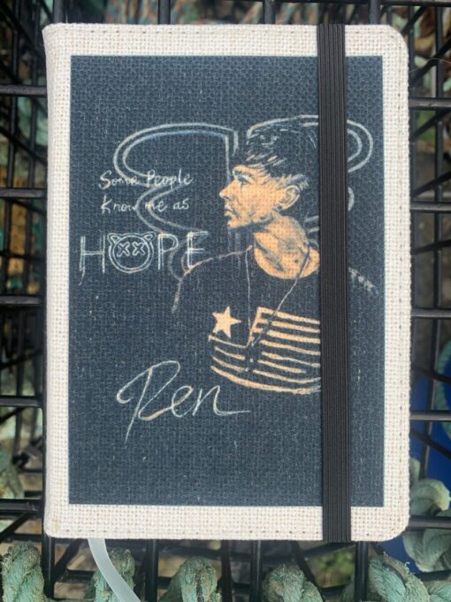 A fabric-covered notebook with a portrait and text: "Some people know me as Hope. Ren." A black elastic band secures it.