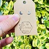 Hand holding tan tag with crescent moon earrings and "the beaded magpie" logo, against variegated green plant background.